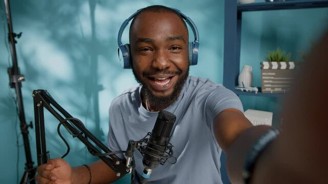 POV of influencer vlogging in studio and talking with microphone to subscribers. Vlogger holding camera to record video for vlog channel, using headphones. Man with podcast gear