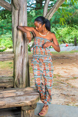 Wearing a strapless fashionable, contemporary style dress top and pants, fashionable sandals, a pretty black woman is standing by an old tree,  raising her arms, looking down, checking her clothes..