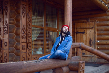 man with a hipster beard in a red knitted hat and a denim jacket is sitting on the stairs at wooden house in the woods