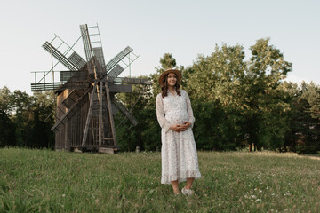 A full-length portrait of a pregnant woman in the windmill background who is holding her belly with her hands on the farm. A happy pregnancy. A lady in a dress and a hat at the meadow in the evening.
