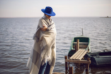 woman in jeans and a sweater and a hat stands on a pier with boats by lake in autumn