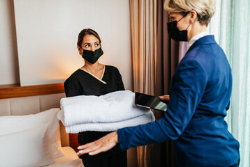 Hotel manager explains to maid how best to tidy up the room. They are both wearing protective face...