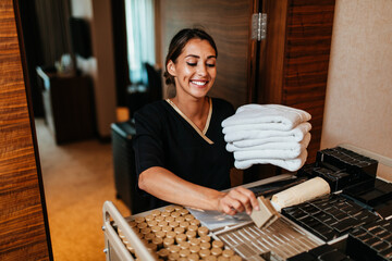 Beautiful young hotel chambermaid in uniform bringing clean towels and other supplies to hotel room. - obrazy, fototapety, plakaty