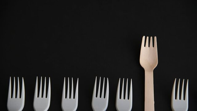 Wooden disposable fork made from birch tree moves up above plastic ones lying on black background. Close-up top view. Copy space for your text. Biodegradable cutlery. Environmental protection theme.