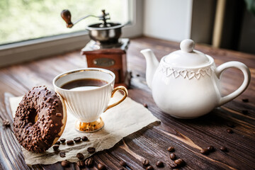 Morning tea with sweet doughnut. A white vintage cup of coffee next to a teapot and freshly ground coffee in an antique coffee grinder