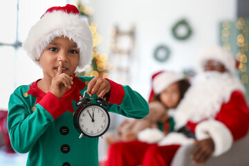Little African-American boy dressed as elf with alarm clock showing silence gesture at home