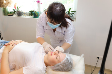 A beautician in sterile white gloves and a white lab coat is leaning over a client and trying to...