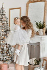 Adult woman with tender makeup and long hair posing with her cute toddler daughter in decorated Christmas interior