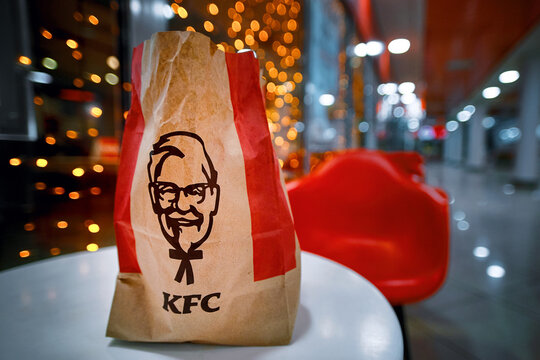 Minsk, Belarus. Jan 2021. KFC paper bag with Kentucky Fried Chicken standing on the table against christmas lights background. KFC paper package and festive Christmas garland in bokeh