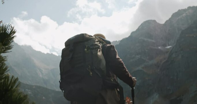 Male hiker backpacker with trekking poles reaches spot overlooking beautiful lake and mountain peaks. Man traveler steps on big stone, stands looking at amazing highlands landscape on sunny day
