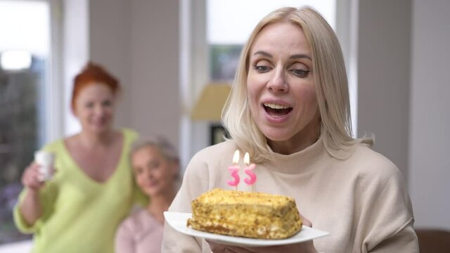 Charming happy woman making wish blowing out candles on 35 birthday looking back at friends toasting and looking at camera smiling. Confident beautiful slim lady posing on holiday at home