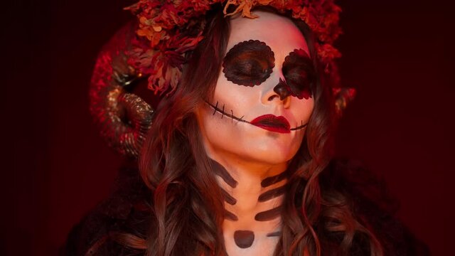 woman in rags halloween witch costume with calavera catrina skull makeup, horns and dried flowers on her head in a dark red room peeks at the camera. Close-up