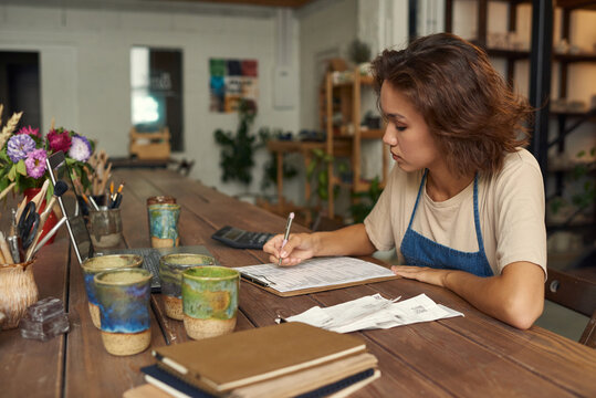 Concentrated young woman sitting at wooden table with ceramic mugs and ananlyzing receipts while working in pottery workshop