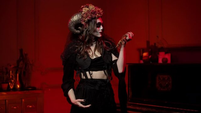 Halloween witch brunette with skull Calavera Katrina makeup, horns and dried flowers dancing with clawed paws of an eagle in a dark room with red light