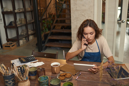 Busy young female potter sitting at desk with tools and ceramic mugs and using tablet while calling supplier in workshop