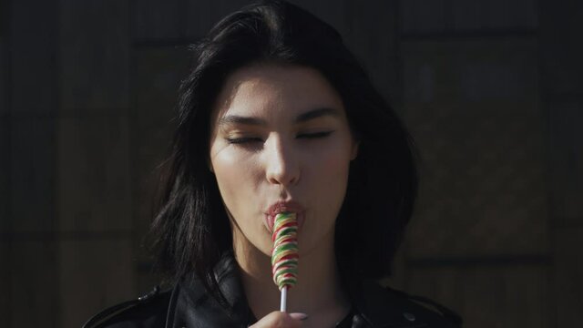 Portrait happy young brunette woman in trendy stylish look wearing black leather jacket seductively licks colorful lollipop candy looking at camera and smiling standing by wall in city street