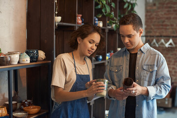 Positive young saleswoman in apron standing against shelves and showing ceramic mug to buyer in...