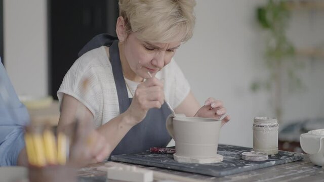 Woman Potter Attaching Handle To Mug In Ceramics Studio. Female potter shaping and carving mug in pottery workshop, studio