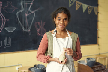 Portrait of smiling African American girl in white apron standing with handmade vase against blackboard at pottery class