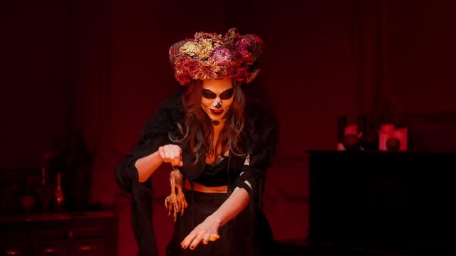 woman in rags halloween witch costume with calavera catrina skull makeup, horns and dried flowers on her head in a dark red room casts a spell over a fire and throws an eagle's paws into a bonfire