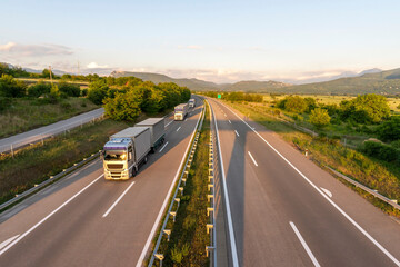 Convoy of Big Transportation trucks departing on a country highway under a sunset sky. 