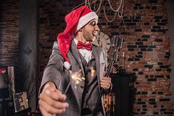 a man in a formal suit and a Christmas hat with a microphone with a sparkler in his hand and a clock in the background
