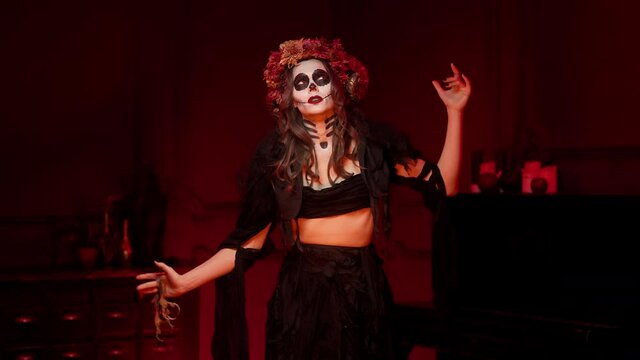 Halloween witch conjures, dances in a trance in the red room by the fire, throws the paws of a bird into the fire, waves her arms, closes and opens her eyes. Day of the Dead horned skull makeup