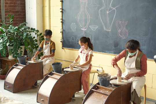 Group of multi-ethnic children in masks and aprons making clay vases using pottery wheels in workshop