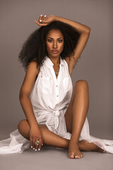 Portrait of a beautiful young Latin woman with long curly hair and beautiful makeup sitting by herself barefoot on the floor inside a studio with a grey background wearing a white dress and jewelry.