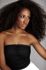 Naklejka premium Closeup portrait of a serene young Latin woman with long curly hair and beautiful makeup posing by herself inside a studio with a grey background wearing a black wrapped top and white shorts.
