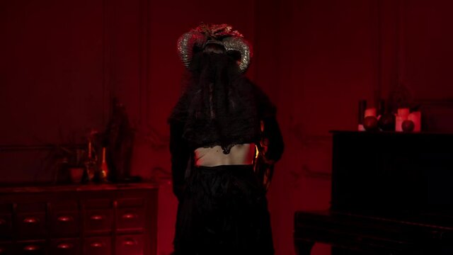 female shaman in form of calavera is turned with her back to camera, conjures over fire, turns to camera, glances at viewer. Makeup, halloween costume in style of day of dead, with horns, in red room
