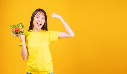Health care food concept. Excited cheerful young woman slim body holding glass bowl with organic vegetables raised own arm for good healthy while standing over isolated yellow background.