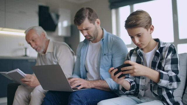 Grandfather reading book, son working on laptop, grandson playing video game