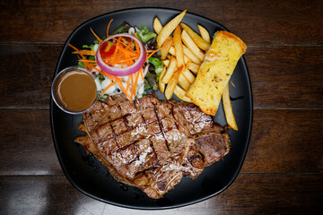 T-bone steak with french fries, salad on a black plate placed on a wooden table top view