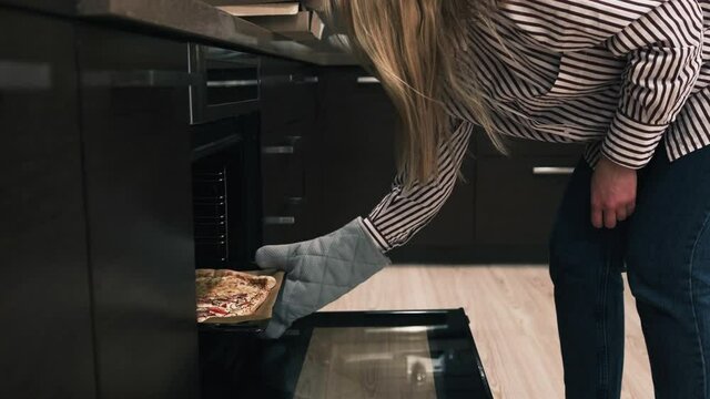 Blonde lady in blue stripped blouse takes pizza out of the oven in dark kitchen. Close up. Slow mo