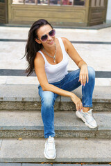 Beautiful caucasian woman with glasses is sitting on the street on a crowded city staircase and smiling. Cute brunette posing in the city while sitting on the steps in casual clothes