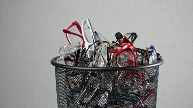 Camera shows pile of multi-colored frame glasses put into black trash bin against grey background as request to improve eyesight closeup
