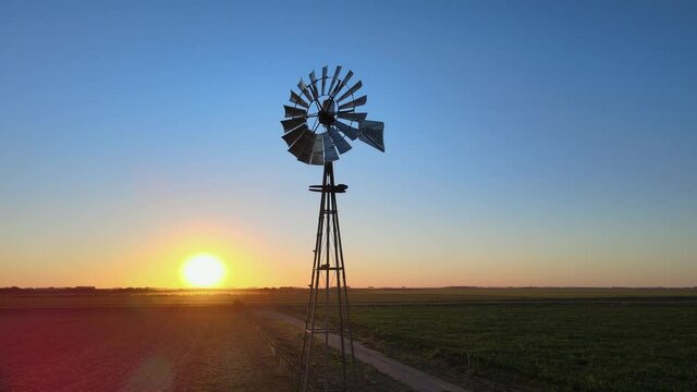 Aerial slow pan shot in open ploughed field with old school cowboy style rustic windmill at the center and sunset in the background, La Pampa, Patagonia, Argentina.