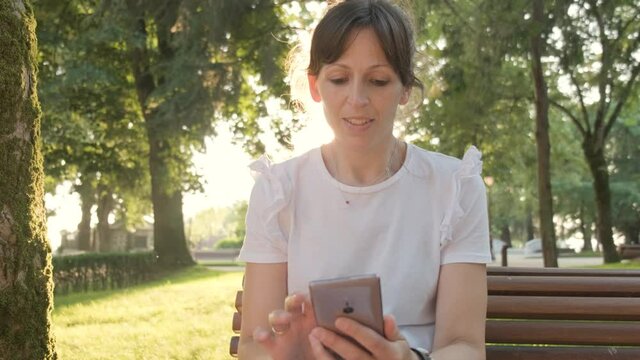 A Woman Scrolling Through Her Phone While Sitting In the Park with Sun Lens Flare Behind Her, Handheld Shot