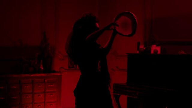 a brunette woman in a suit and make-up of a calavera skeleton with horns and flowers for halloween dance in a dark room with a red light, hits a tambourine, pronounces a spell