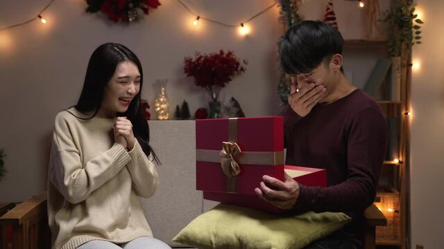 family holiday celebration lifestyle concept. Smiling woman surprising boyfriend with present at home on sofa. shocked young man open mouth and cover with hand while looking bright inside gift