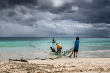 Fishermen pull nets from the sea on a beach in the tropics with dramatic sky on Mahe Island,...