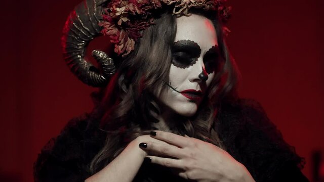 Demanic woman witch on All Saints Day dances in a dark room with red lighting under flashes of lightning, looks at the camera. Makeup, halloween costume in Catrina calavera style. Close-up