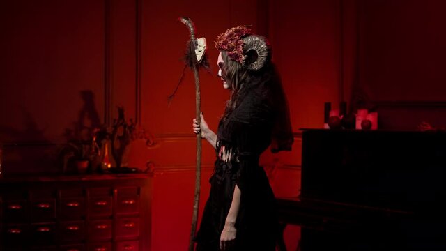 a voodoo woman walks with a staff with a skull through a dark fiery room, turns to face the camera, looks. Halloween skeleton style witch image with horns, sewn mouth