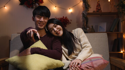 sweet asian young couple in love face camera at Christmas eve enjoy watch tv while sitting under...