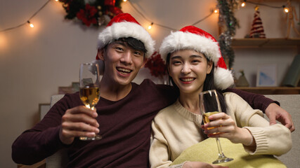 love romance holiday celebration concept. engaged couple with red santa hat holding and drinking wine glasses sitting on sofa during video call online. joy female and male toasting with white wine