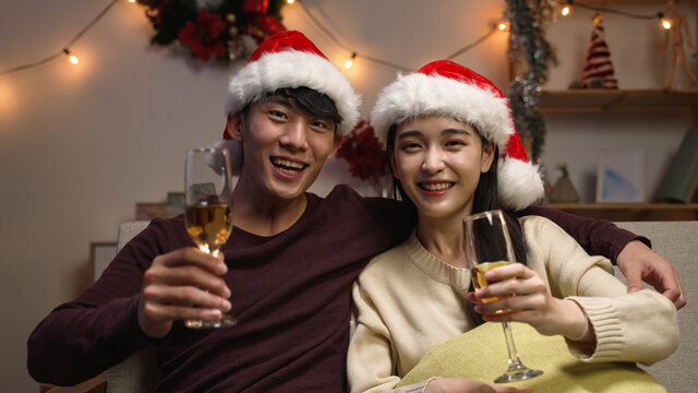 love romance holiday celebration concept. engaged couple with red santa hat holding and drinking wine glasses sitting on sofa during video call online. joy female and male toasting with white wine - Powered by Adobe