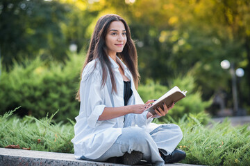 A young and beautiful Arab or Caucasian girl dressed in casual style is reading a book while sitting in the park. Knowledge and education concept.