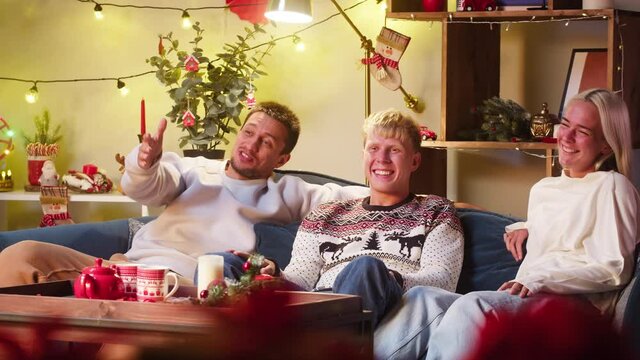 Friends watching television, comedy film. Young people sitting on sofa, watch christmas movie on tv, laughing. Men and women during New Year time at home. Cozy winter evening concept, holidays. 