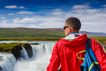 Man in red jacket standing on the rock and looking at Godafoss waterfall. Beautiful landscape in Iceland